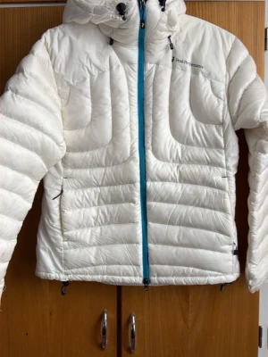 Vit dunjacka från Peak Performance - Snygg vit dunjacka från Peak Performance med blå dragkedja och justerbar huva. Jackan har två sidofickor med dragkedja och diskret logga på bröstet. Perfekt för kalla dagar och riktigt lätt att bära. Klassisk quiltad design och modern passform.