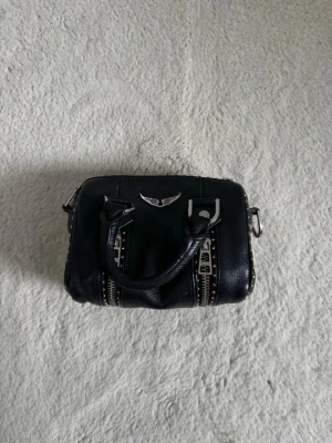 Svart handväska med silverdetaljer - Cool svart handväska i skinn med två rejäla handtag, silverfärgade dragkedjor och nitar längs sidorna. Väskan har en metallfärgad logga framtill och detaljer i metall som ger en edgy vibe. Perfekt storlek för det viktigaste och passar till många outfits. Den är från Zadig Voltaire, ordinarie pris är 2700kr