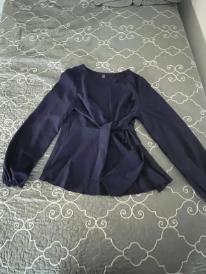 Mörkblå blus med knytband - Stilren mörkblå blus från H&M med långa ärmar och ett snyggt knytband i midjan. Blusen har rund halsringning och är tillverkad i ett lätt, mjukt tyg som ger ett elegant fall. Perfekt för att lyfta vilken outfit som helst.