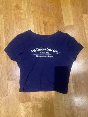 Mörkblå croppad t-shirt Wellness Society - Snygg mörkblå croppad t-shirt från Wellness Society med vit text framtill. Klassisk rund halsringning och korta ärmar. Perfekt för en avslappnad och sportig stil. Tillverkad i mjuk bomull för skön känsla.