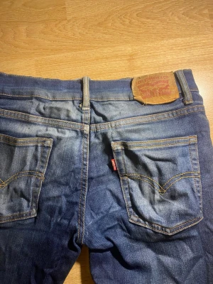 Levi's blå slim taper jeans - Säljer ett par klassiska blå jeans från Levi's i modellen Slim Taper. Jeansen har fem fickor, gul kontrastsöm och en ikonisk läderpatch bak i midjan. Tillverkade i slitstark denim med snygg tvätt och subtila slitningar för en avslappnad look.