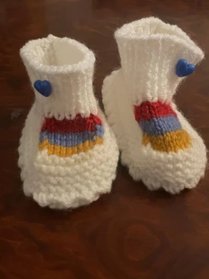 Stickade vita tofflor med hjärtdetalj - Säljer ett par ny handstickade vita tofflor med färgglada ränder i rött, blått och gult framtill. Tofflorna har en blå hjärtformad knapp på sidan och är gjorda i mjukt garn. Perfekta för att hålla fötterna varma och mysiga.