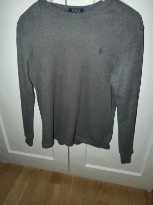 Grå långärmad tröja Polo Ralph Lauren - Snygg grå långärmad tröja från Polo Ralph Lauren med diskret broderad logga på bröstet. Tröjan har rund halsringning och är tillverkad i ett mjukt, stickat material som känns skönt mot huden. Perfekt för en stilren och avslappnad look.