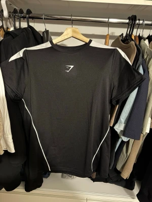 Gymshark Critical T-Shirt - Snygg svart t-shirt från Gymshark med vita paneler på axlarna och vita linjer längs sidorna. Tillverkad i ett lätt och andningsbart material som passar perfekt till träning. Gymshark-logga på bröstet och i nacken. Modellen är slim fit och storlek är medium.
