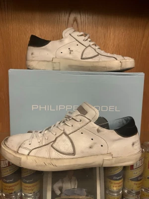 Vita Philippe Model sneakers med svart häl - Snygga vita sneakers från Philippe Model med svarta detaljer på hälen och gråa inslag runt sulan. Klassisk låg modell med vita skosnören och ikonisk sköld-logga på sidan. Perfekta för en trendig och avslappnad streetstyle.