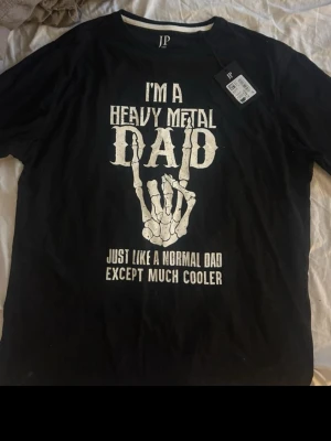 Svart t-shirt Heavy Metal Dad JP1880 - Cool svart t-shirt från JP1880 med trycket 'I'm a Heavy Metal Dad' och ett skelett-handmotiv på bröstet. T-shirten har rund hals och korta ärmar. Perfekt för dig som gillar rockig stil och vill sticka ut lite extra.