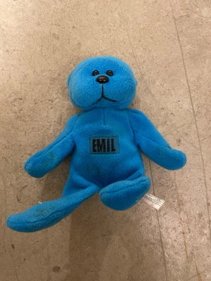 Blå mjukisdjur nalle med text EMIL - Söt blå mjukisdjur nalle i plysch med broderad svart text 'EMIL' på magen. Nallen har svarta plastögon och nos samt mjuka, böjbara armar och ben. Perfekt som personlig present eller dekoration.