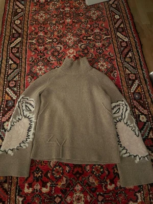 Beige stickad tröja Zadig & Voltaire - Cool beige stickad tröja från Zadig & Voltaire med hög krage och unika grafiska mönster i vitt och svart på ärmarna. Köpte den i Miami och har bara använt 1 gång. Den är perfekt för dig som vill ha något annorlunda och trendigt i garderoben. 