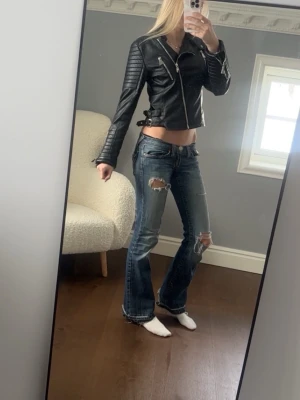 Svart 2000s skinnjacka med detaljer - Snygg svart skinnjacka med silvriga dragkedjor och quiltade partier på ärmarna. Jackan har justerbara spännen i sidorna och en kort, figurnära passform. Perfekt för dig som vill ha en edgy look.