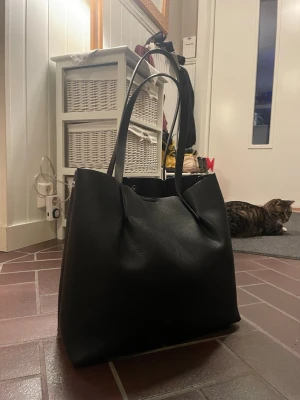 Stor svart handväska  - Snygg och rymlig svart handväska i slätt konstläder med två långa axelband. 