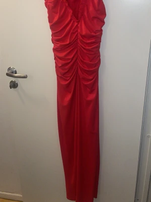 Röd långklänning med rynkade detaljer - Säljer en snygg röd långklänning med rynkade detaljer framtill. Klänningen har en figurnära passform och är ärmlös, perfekt för dig som vill sticka ut. Materialet känns mjukt och följsamt, troligen polyester. Perfekt till speciella tillfällen!