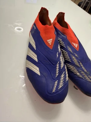 Adidas Predator blå/röda fotbollsskor - Säljer ett par Adidas Predator fotbollsskor i blått med vita detaljer och röd strumpliknande krage. Skorna har en modern, strömlinjeformad design utan snörning och är tillverkade i syntetmaterial för bästa bollkontroll. Perfekta för dig som vill sticka ut på planen!