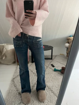 Blå bootcut jeans med fickdetaljer - Säljer ett par blå bootcut jeans med snygga fickdetaljer framtill och bak. Jeansen har låg midja och klassisk denimlook med kontrastsömmar. Perfekta för dig som gillar en avslappnad men trendig stil.