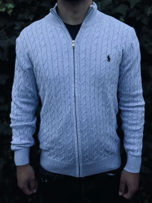 Ljusblå kabelstickad ziptröja Polo Ralph Lauren - Snygg ljusblå kabelstickad ziptröja från Polo Ralph Lauren med broderad logga på bröstet. Tröjan har hel dragkedja, ribbade muddar och krage. Perfekt för lager-på-lager och ger en klassisk look.