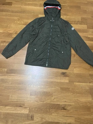 Oliv grön Moncler vindjacka  - Säljer en oliv grön vindjacka från Moncler med huva och dragkedja framtill. Jackan har Moncler-logga på ärmen, två fickor med knappar och snygg detalj med röd, vit och blå rand på huvan. Tillverkad i lätt polyester, perfekt för blåsiga dagar. Storlek 4 som är ungefär L. Helt ny 