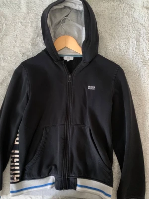 Svart zip hoodie från BOSS - Snygg svart hoodie med dragkedja från BOSS. Har vit logga på bröstet, huva med grå insida och fickor framtill. Ribbad mudd med blå och vit rand längst ner samt tryck på ena ärmen. Perfekt för en chill och stilren look.