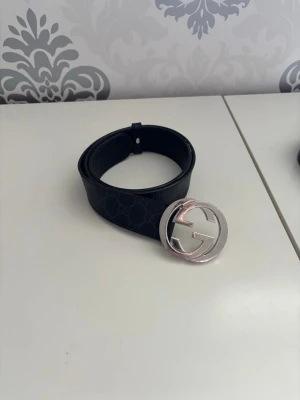Svart Gucci bälte med silverdetalj - Snyggt svart bälte från Gucci med klassiskt GG-mönster och stor rund silverspänne. Bältet är gjort i skinn. Äkta bälte men har ej något kvar som kan bevisa det, där av priset. Nytt pris på bältet är 4800kr men mitt pris är 850kr