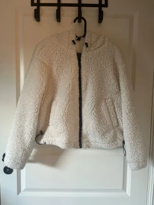 Vit teddyjacka från H&M Divided - Supermjuk och fluffig vit teddyjacka från H&M Divided. Jackan har dragkedja framtill, två fickor och justerbar dragsko i nederkant. Perfekt för kyliga dagar när du vill vara både varm och snygg.