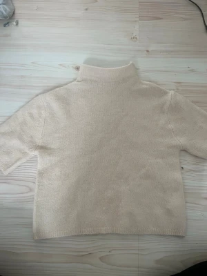 Beige stickad polotröja - En enkel och stilren beige stickad polotröja med korta ärmar och hög krage. Tröjan är gjord i mjukt material och har en rak passform som är lätt att matcha med olika outfits. Perfekt för lager-på-lager och en clean look.