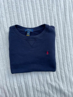 Mörkblå sweatshirt från Polo Ralph Lauren - Klassisk mörkblå sweatshirt från Polo Ralph Lauren med rund halsringning och den ikoniska röda logotypen broderad på bröstet. Tröjan har ribbade muddar vid ärmslut och nederkant. Perfekt för en avslappnad och stilren look.