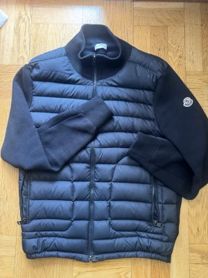 Moncler cardigan - Snygg svart cardiganjacka från Moncler med quiltad framsida och stickade ärmar. Jackan har dragkedja framtill, ribbad krage och muddar samt två fickor med dragkedja. Moncler-logga på ena ärmen. Perfekt för lager-på-lager och streetstyle.