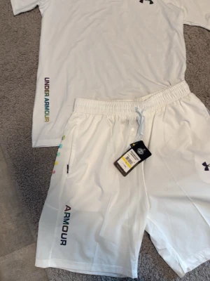 Vita shorts från Under Armour - Snygga vita shorts från Under Armour med färgglad logga längs benet. Shortsen har elastisk midja med snörning och sidofickor. Perfekta för träning eller chill dagar. Tillverkade i ett lätt och snabbtorkande material.