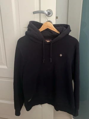 Morris Hoodie - Storlek S. Snygg mörkblå hoodie med huva och dragsnören. På bröstet finns en liten broderad detalj. Hoodien har en klassisk passform och är tillverkad i mjukt material som känns skönt mot huden. Perfekt för en avslappnad och stilren look.  Hoodien är i nyskick och nypriset ligger på 1500kr.