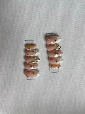 Press-on naglar med guld och tigerdetaljer - Snygga press-on naglar i mandelform med ljusrosa bas, vita tippar och dekorerade med guldfärgade pärlor. Några naglar har coola tigerinspirerade detaljer i svart och orange. Perfekt för dig som vill ha unika och trendiga naglar.