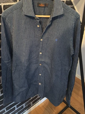 Randig blå skjorta från Morris - Snygg blå skjorta från Workers med vita smala ränder och klassisk krage. Skjortan har långa ärmar, knappar framtill och en liten broderad logga på bröstet. Perfekt för dig som gillar stilrena detaljer och tidlös design.
