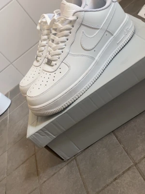 Nike Air Force 1 vita sneakers - Klassiska Nike Air Force 1 sneakers i helvitt läder med perforerad tå, vit sula och ikonisk swoosh på sidorna. Skorna har lågt skaft, vita skosnören och en chunky sula som ger en clean och tidlös look.