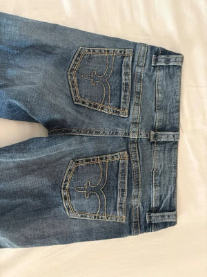 Blå bootcut jeans med broderade fickor - Säljer ett par blå bootcut jeans med snygga broderade bakfickor och slitningar framtill. Jeansen har låg midja, klassisk femficksmodell och är tillverkade i mjuk denim. Perfekta för dig som gillar retrovibbar och vill ha ett par jeans med unik detalj på fickorna.