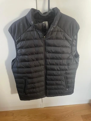 Svart dunväst från Decathlon - Snygg svart dunväst från Decathlon med quiltad design och dragkedja framtill. Västen har två praktiska fickor med dragkedja och hög krage för extra värme. Perfekt för lager-på-lager och enkel att matcha med olika outfits.