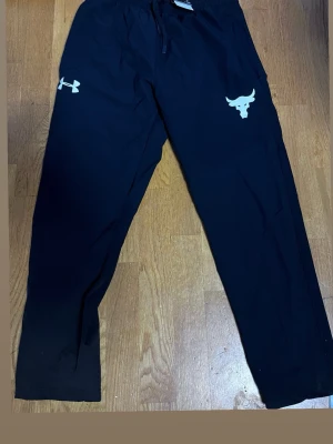 Svarta vindbyxor Under Armour - Svarta vindbyxor från Under Armour med bulls-logga och dragsko i midjan. Byxorna har raka ben och resår i midjan. Perfekta för chill, dåligt väder  eller träning.
