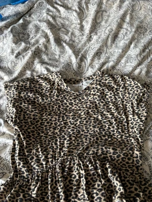 Leopardmönstrad kort klänning - Kort klänning med leopardmönster i beige och svart. Klänningen har korta ärmar, v-ringning och en lätt rynkad midja för en avslappnad passform. Perfekt för dig som vill sticka ut med ett djurmönster.