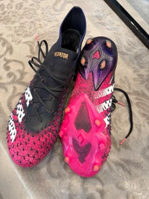 Adidas Predator rosa/svarta fotbollsskor - Säljer ett par Adidas Predator fotbollsskor i svart och rosa med vita detaljer och klassiska tre ränder på sidan. Skorna har hög krage, snörning och gummidetaljer på ovandelen för extra grepp. Sulan är i rosa med fasta dobbar, perfekt för spel på gräs.
