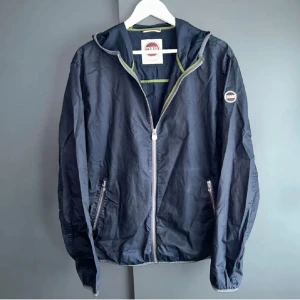 Colmar windbraker - Svart vindjacka från Colmar med huva och hel dragkedja framtill. Jackan har två sidofickor med dragkedja och en diskret logotyp på vänster ärm. Lätt och smidig design med gröna detaljer på insidan. Perfekt för dig som gillar stilren och sportig look. Mycket bra skick inga skavanker eller hål. Storlek 52 dvs L men tycker den sitter mer likt m.