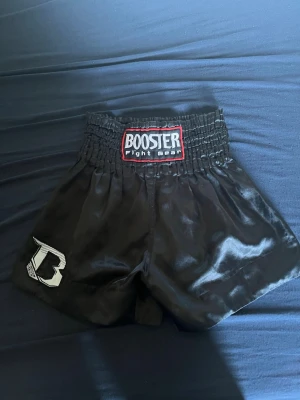 Svarta thaishorts Booster Fight Gear - Säljer ett par svarta thaishorts från Booster Fight Gear med bred resår i midjan och vit B-logga på ena benet. Shortsen är tillverkade i glansigt material och har en klassisk passform för kampsport.