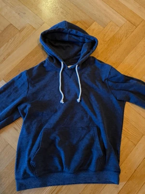 Mörkblå hoodie med vita snören - En stilren mörkblå hoodie med klassisk känguruficka och vita dragsnören i huvan. Hoodien har en avslappnad passform och ribbade muddar vid ärmar och nederkant. Perfekt för en chill och casual look.