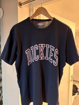Marinblå Dickies t-shirt - Snygg marinblå t-shirt från Dickies med stort vitt och rött tryck på bröstet. Klassisk passform och rund halsringning. Perfekt till jeans eller shorts för en avslappnad look.