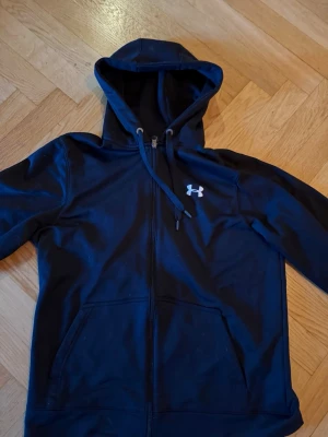 Svart hoodie från Under Armour - Snygg  hoodie från Under Armour med dragkedja framtill och två stora fickor. Klassisk logga på bröstet och justerbar huva med snören. Perfekt för chill eller träning. Tillverkad i mjukt och bekvämt material.