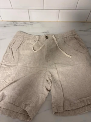Beige linneshorts från H&M - Snygga beige shorts från H&M i linne med regular fit. De har elastisk midja med snörning, knappstängning och två sidofickor. Perfekta för varma dagar och ger en avslappnad vibe.