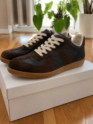Maison Margiela Gats brun/svart - Snygga sneakers från Maison Margiela i brunt mocka och svart läder med vita snören och gummisula. Klassisk low-top siluett som passar perfekt till streetwear och casual outfits. Diskret branding och stilren design.