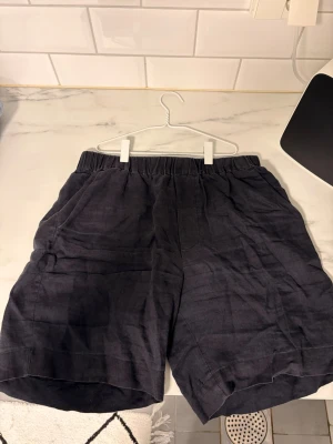 Mörkblå Sandö linne shorts - Säljer ett par mörkblå linne shorts med elastisk midja och avslappnad passform. Shortsen har sidofickor fram och en bakficka. Perfekta för en chill och stilren look under varmare dagar.
