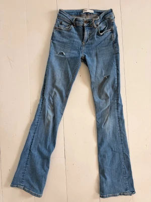 Blå bootcut jeans från ZARA - Säljer ett par ljusblå bootcut jeans från ZARA med slitningar på låret och knät. Jeansen har klassisk femficksmodell, hög midja och är tillverkade i denim med lätt stretch. Perfekta för dig som gillar en avslappnad men trendig look.