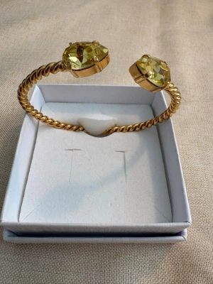 Caroline Svedbom Armand i toppskick - Guldigt armband med stora stenar - Snyggt armband i guldton med tvinnad design och två stora, facetterade gula stenar i ändarna. Armbandet är öppet och justerbart, perfekt för att ge en lyxig touch till din outfit. Stenarna fångar ljuset och ger extra bling.