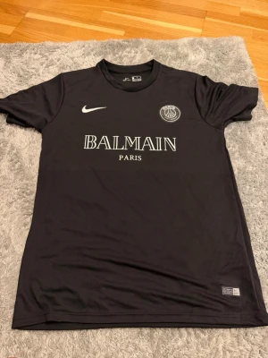 Svart PSG x Balmain t-shirt Nike - Snygg svart t-shirt från Nike i samarbete med PSG och Balmain Paris. Klassisk rund hals, korta ärmar och tryckta loggor framtill. Perfekt för dig som gillar streetwear och sportig stil.