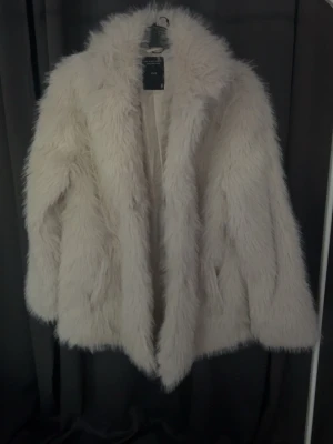 Vit fluffig fuskpälsjacka - Säljer en supermjuk och fluffig vit fuskpälsjacka med bred krage och öppen front. Jackan har en oversized passform och är perfekt för att hålla värmen under kalla dagar. 