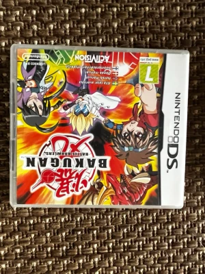 Bakugan Battle Brawlers till Nintendo DS - Bakugan Battle Brawlers är ett actionfyllt spel till Nintendo DS med färgstark animegrafik på omslaget. Spelet har en vit plastbox och motiv med karaktärer och drakar i rött, gult och svart. Perfekt för dig som gillar strategi och äventyr i spelvärlden.