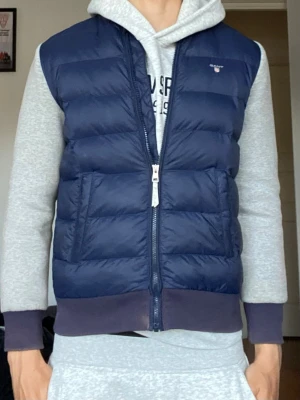 Marinblå dunväst från Gant - Snygg marinblå dunväst från Gant med quiltad design och två stora fickor framtill. Västen har en hög krage och dragkedja hela vägen. Perfekt att slänga över en hoodie för en chill och sportig look. Klassisk Gant-logga på bröstet.