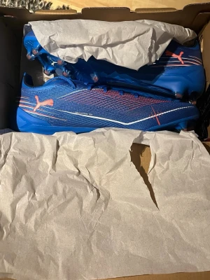 Blåa Puma fotbollsskor med dobbar - Säljer ett par snygga blåa fotbollsskor från Puma med rosa detaljer och vita linjer. Skorna har låg profil, snörning och slitstark syntetisk ovandel. Dobbarna är utformade för bra grepp på planen och skon har en sportig, modern look.